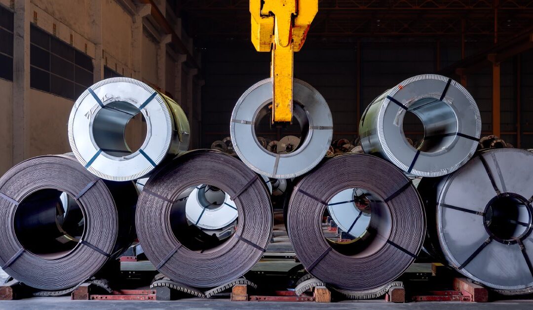 Several rolls of large steel coils in a warehouse center. A yellow crane operates above the organized materials.